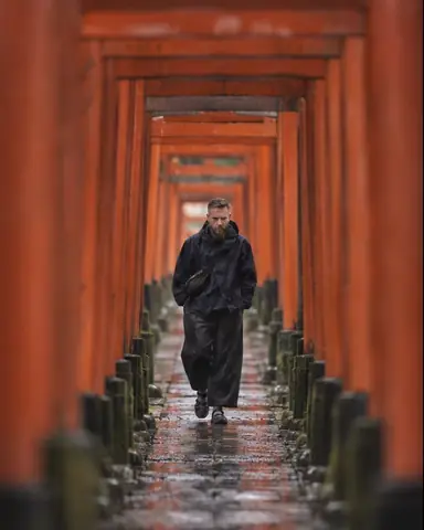 A bearded man stands deep inside a tunnel of vermilion torii gates, a dozen soft out-of-focus gates framing him