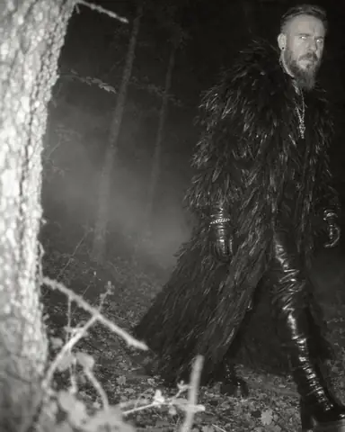 Grainy night-vision view of a bearded man striding past a camera trap, half out of frame, eyes catching the flash