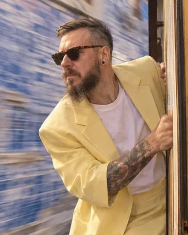 A bearded man in a pastel-yellow suit holds the pole on a Lisbon tram step, the street streaking behind
