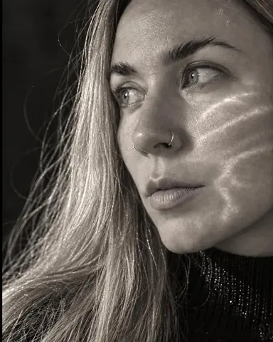 Close three-quarter view of a woman with long blond hair at the frame edge, a ripple of refracted light across her cheekbone