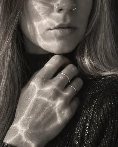 A woman with long blond hair raises a ringed hand to her black high neck, bright bands of refracted light on her knuckles