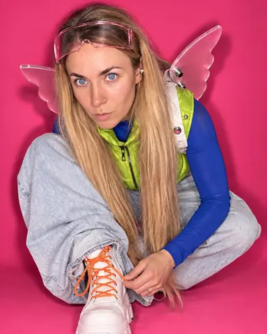 A woman with long blond hair crouches near the lens reaching for an orange boot lace, ring catchlights in both eyes