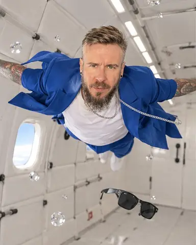 A bearded man floats horizontally across a white padded cabin, arms spread, his blue suit jacket billowing open