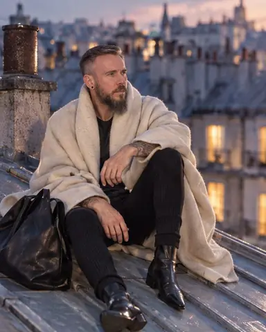 A bearded man sits on a sloped zinc roof in Paris at blue hour, chimneys around him and windows glowing warm