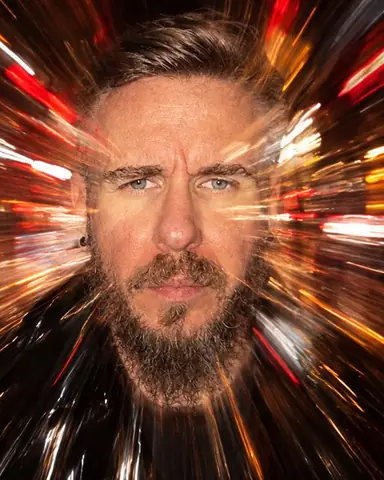 A bearded man's face at the center of radial light streaks bursting outward, half of it frozen by flash