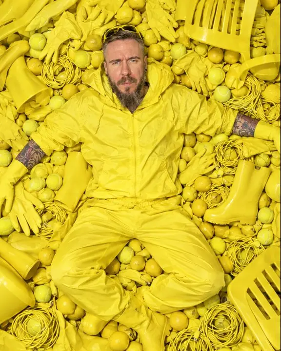 Overhead view of a bearded man lying on his back in a pile of acid-yellow objects, arms spread, looking up