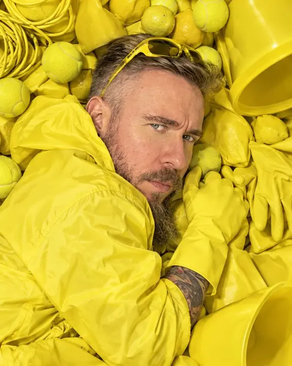 A man curled on his side in a yellow pile, only his face, one shoulder and a gloved hand showing among the objects