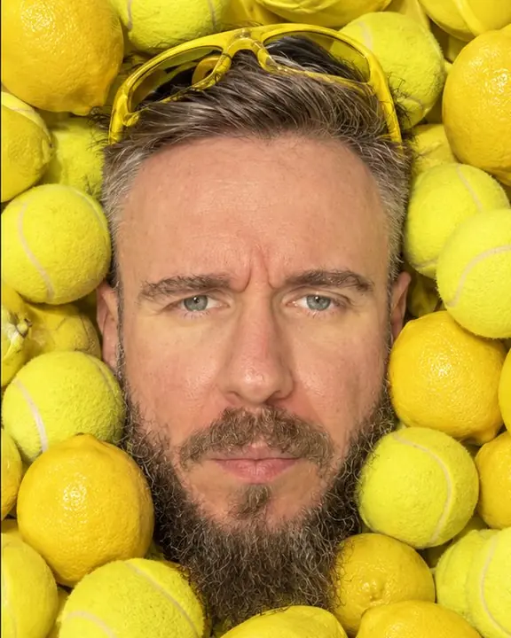 Close overhead view of a bearded face pressed between yellow tennis balls and lemons, eyes to the lens