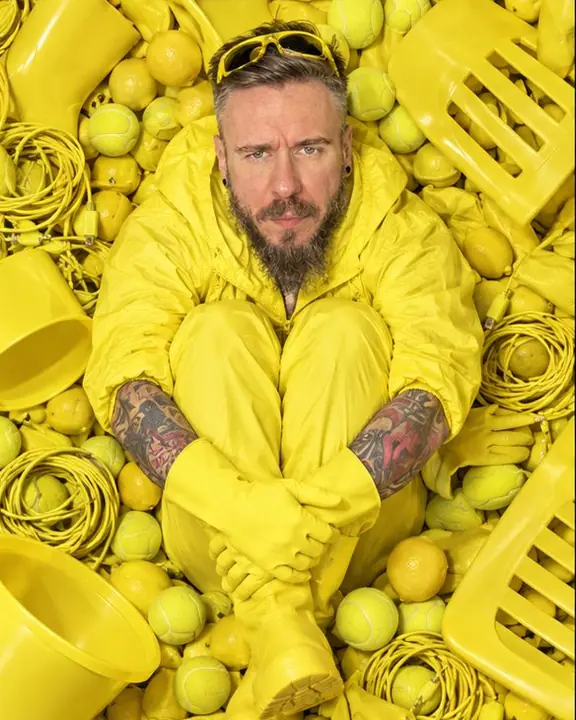 A man in an acid-yellow tracksuit sits up in the pile hugging his knees, objects up to his chest, looking up