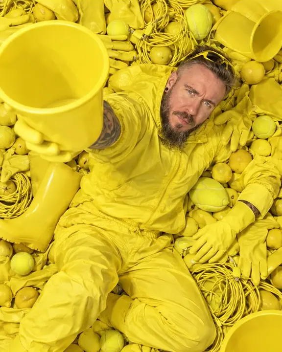 A bearded man lies diagonally across a sea of yellow objects, one arm raised holding a yellow plastic bucket
