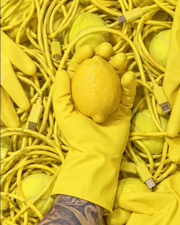 A yellow-gloved hand holds a lemon on top of tangled yellow cables and rubber gloves, seen from above