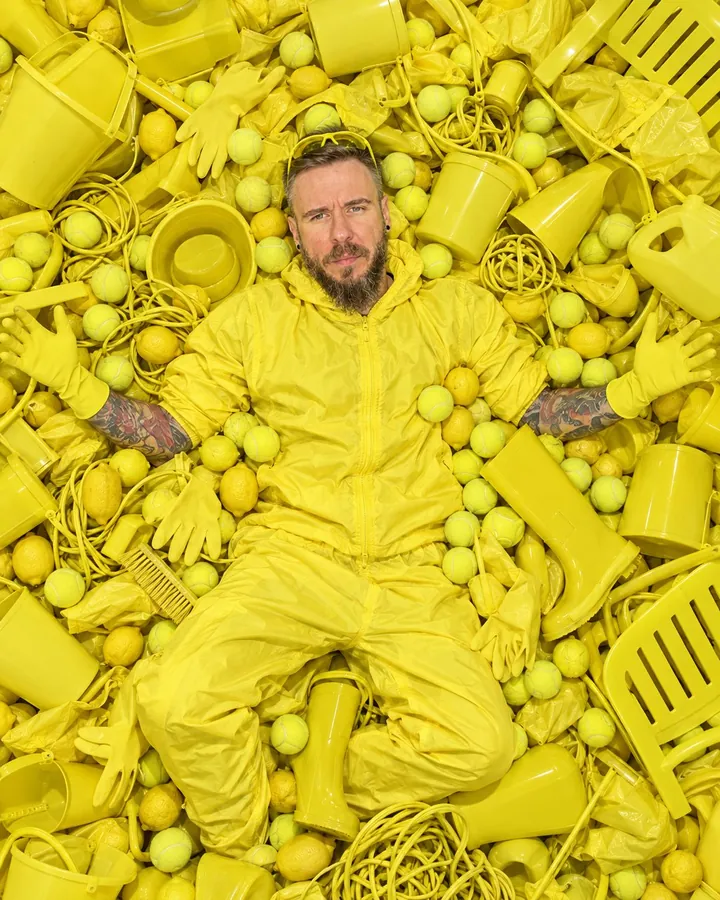 Overhead view of a bearded man lying on his back in a pile of acid-yellow objects, arms spread, looking up