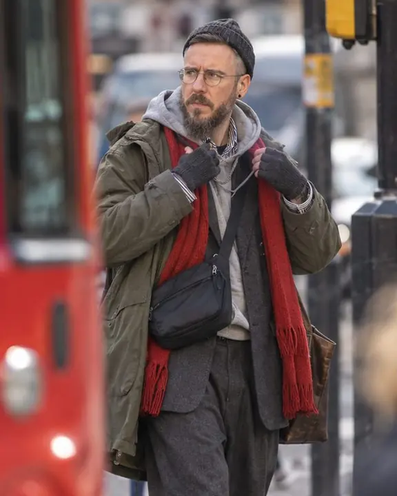 A man pauses at a street corner adjusting a long red scarf, glancing sideways, a blurred red bus at the left edge
