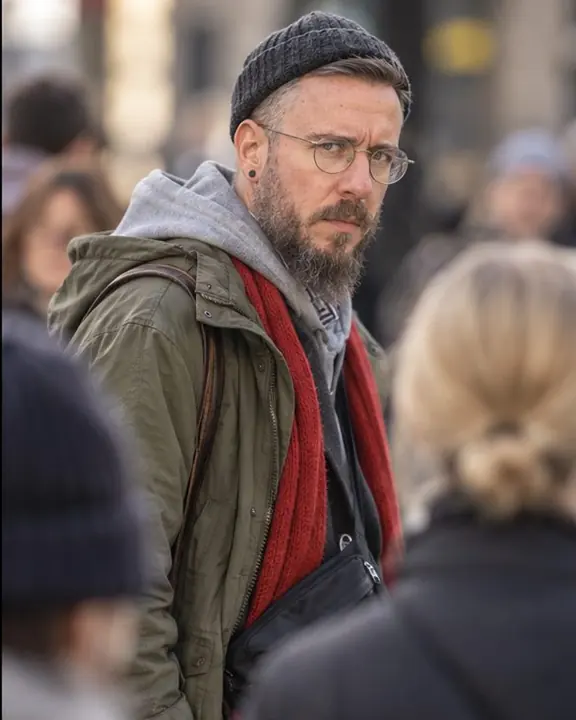 Waist-up view through a gap in a crowd as a bearded man in a parka and blazer turns his head toward the camera