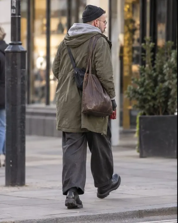A man walks away along a sidewalk, seen from behind and to the side, bag straps and stacked layers against shop windows