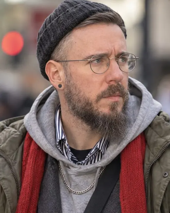 Tight portrait of a bearded man in a beanie and glasses at a crossing, looking aside, soft city signs layered behind