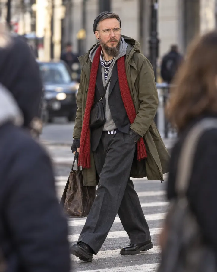 A bearded man in a layered olive parka strides across a crosswalk between two blurred pedestrians in the foreground