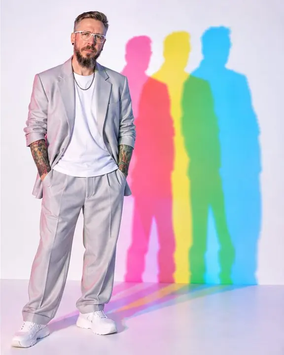 A bearded man in a pale silver-gray suit stands left of center, three cyan, magenta and yellow shadows on the wall