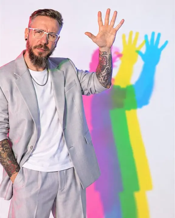 A man raises one hand with fingers spread, its three colored shadows overlapping on a bright white wall