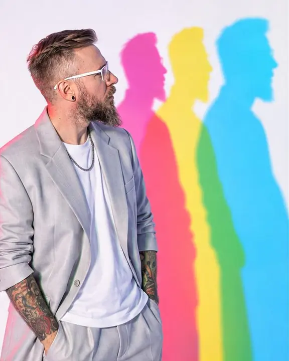 Waist-up profile of a bearded man in white-framed glasses turned to study his own colored shadows with curiosity