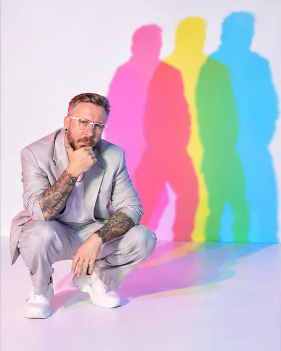 A man crouches low on a white floor in a boxy pale suit, cyan, magenta and yellow shadows stretching up the wall