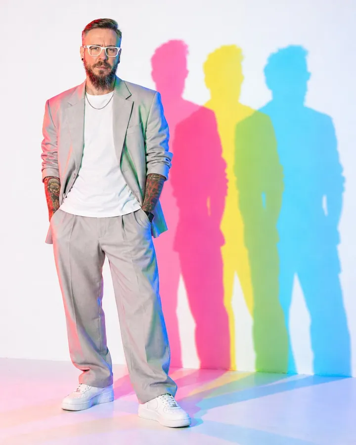 A bearded man in a pale silver-gray suit stands left of center, three cyan, magenta and yellow shadows on the wall