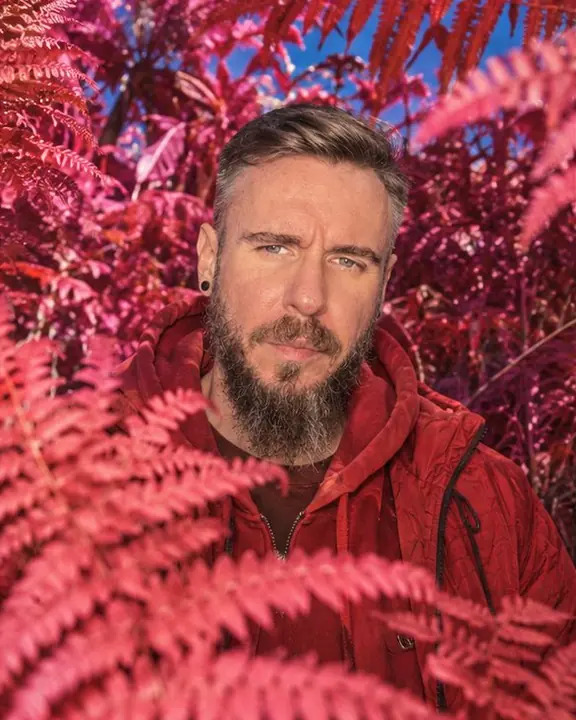 Waist-up of a bearded man half hidden behind scarlet fern fronds, looking at the lens in infrared color