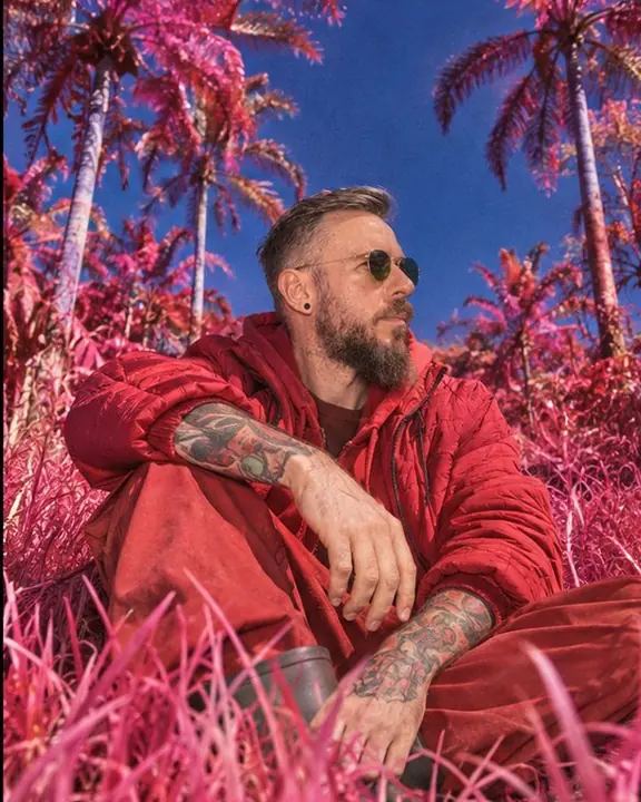 Low-angle view of a man sitting in tall pink grass, palm trees against a deep cobalt sky above him