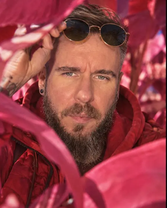 Close portrait of a bearded man framed by magenta leaves, green-tinted round sunglasses pushed up