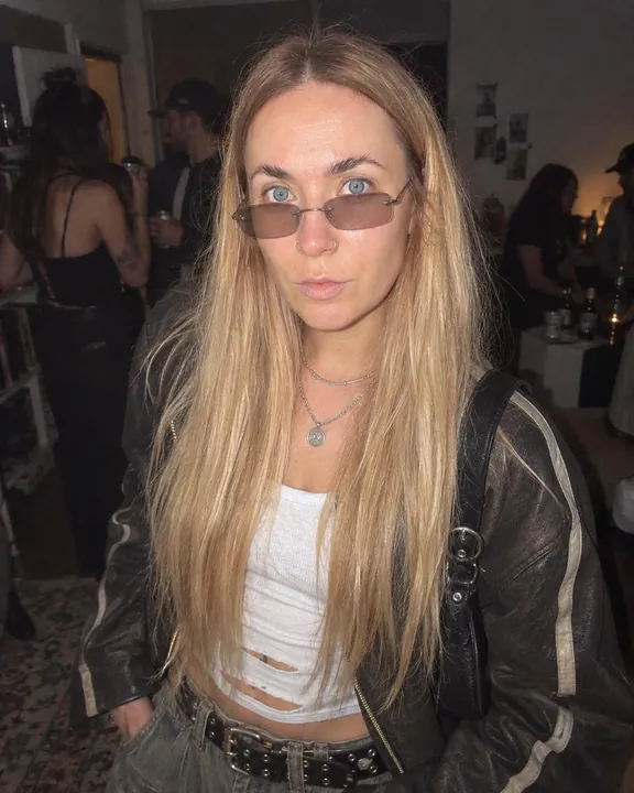 A woman with long blond hair in hard flash, tinted rimless glasses low on her nose, worn leather jacket, guests behind