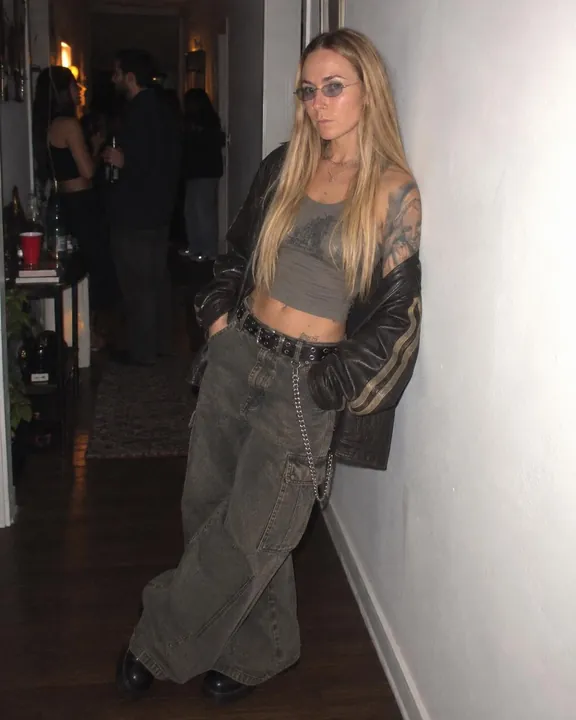 A woman with long blond hair leans on a white hallway wall, leather jacket off one shoulder, wide jeans with a wallet chain