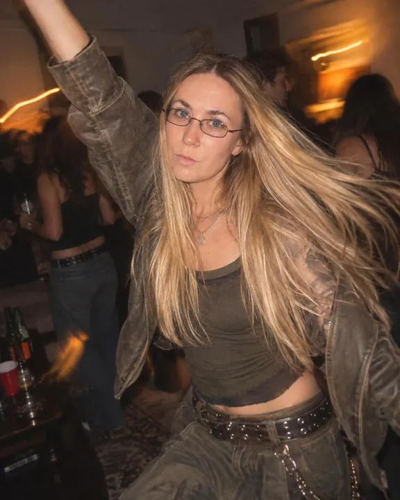 A woman with long blond hair dances with one arm up, hair swinging, thin glasses on, warm lamp streaks behind her