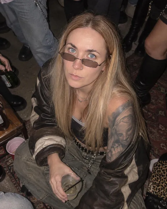 From above, a woman with long blond hair sits on a rug among guests' legs, tinted glasses on, a plastic cup in hand