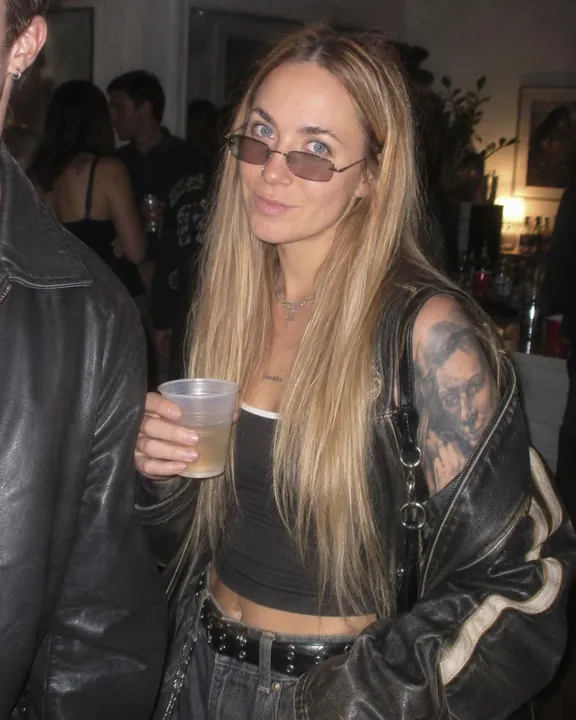 A woman with long blond hair smiles with a plastic cup in hand, tinted glasses on, a man's leather shoulder at the frame edge
