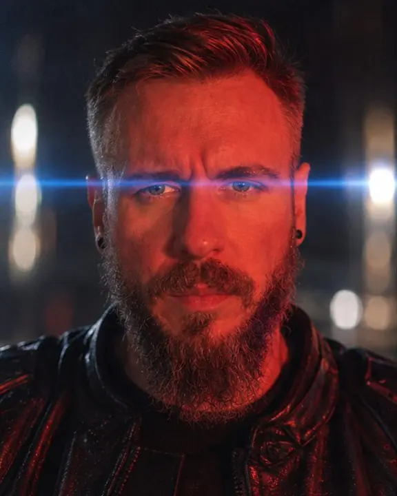 Head and shoulders of a bearded man lit red, a thin blue horizontal flare crossing the frame exactly at eye level