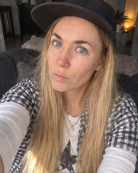 High-angle selfie of a woman with long blond hair on a sofa, head tilted, open gingham shirt and silver chain in window light