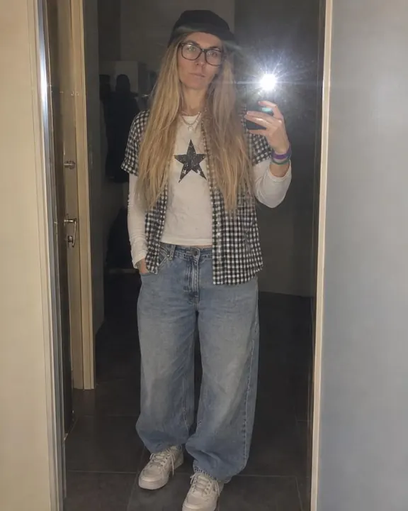 Hallway mirror selfie of a woman with long blond hair in a flat-brim cap, phone at chest height, LED flash flaring white
