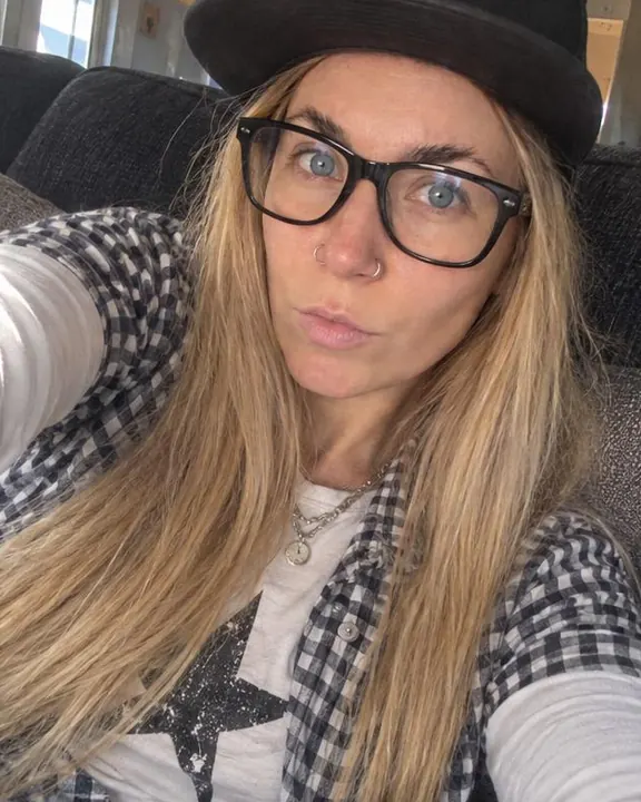 A woman with long blond hair slouches in sofa cushions holding a phone out, big black glasses, the crop cutting her cap