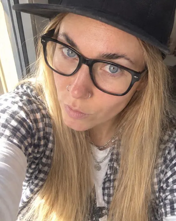 Close diagonal selfie of a woman with long blond hair by a window, chin down, eyes up, sunlight slightly blown on her face