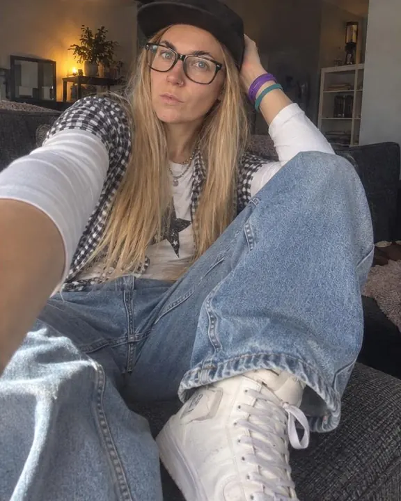 A woman with long blond hair sits on a sofa arm with the phone low and angled up, wide jeans and white high-tops in frame
