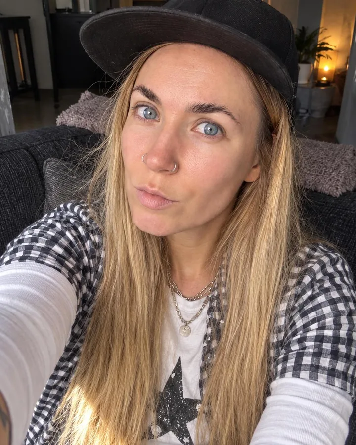 High-angle selfie of a woman with long blond hair on a sofa, head tilted, open gingham shirt and silver chain in window light