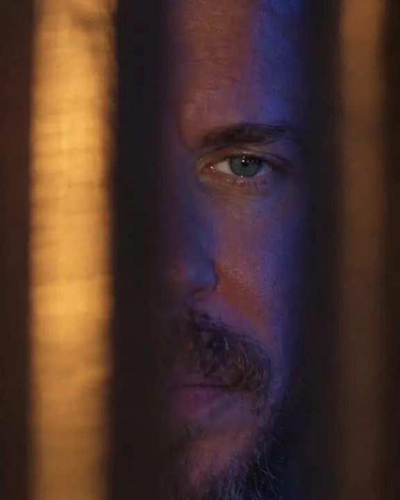 Close-up of one eye of a bearded man seen through a narrow gap between soft bamboo slats