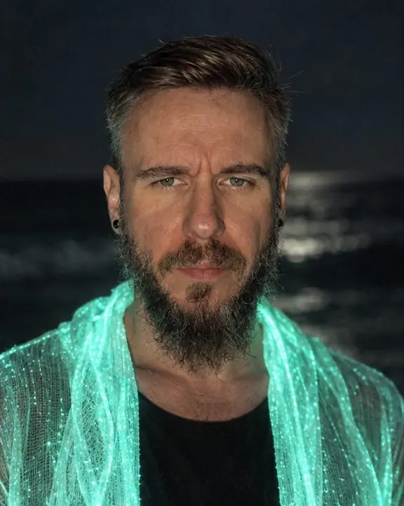 Waist-up shot of a bearded man whose face is lit from below by a softly glowing collar, eyes catching light
