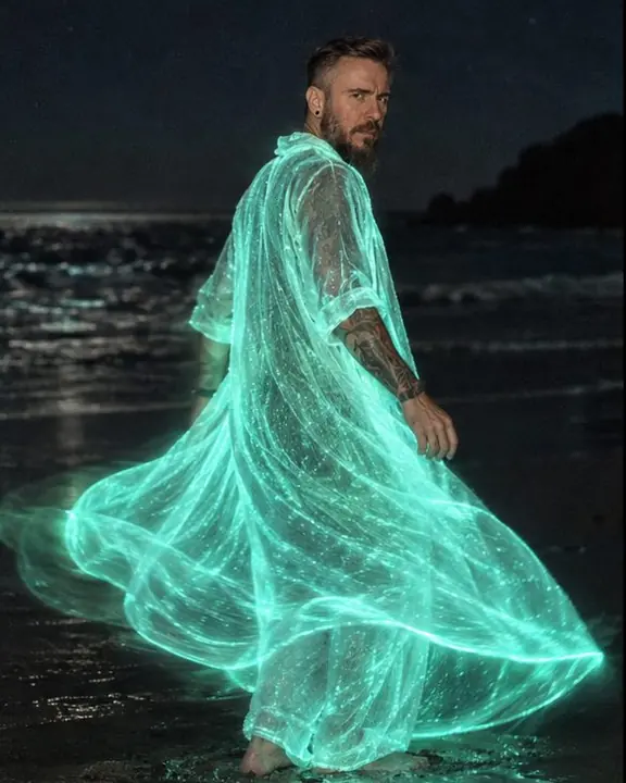 A man turns slowly on a dark beach as the hem of his glowing coat leaves a soft cyan arc of light