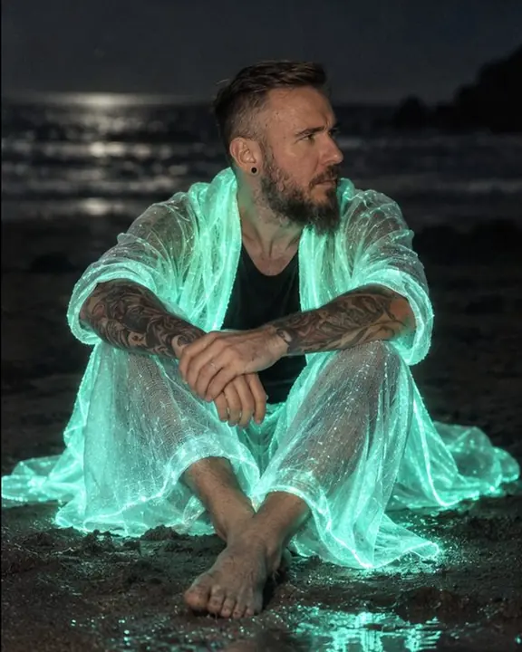 A bearded man sits in the sand with knees up, glowing blue-green fabric pooled around him in the dark