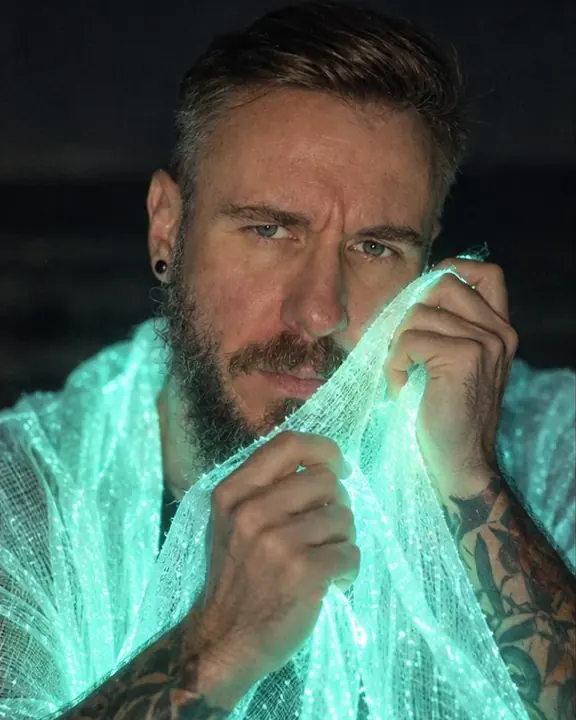 Close shot of a bearded man holding glowing fabric near his face on a night beach, soft cyan light on skin