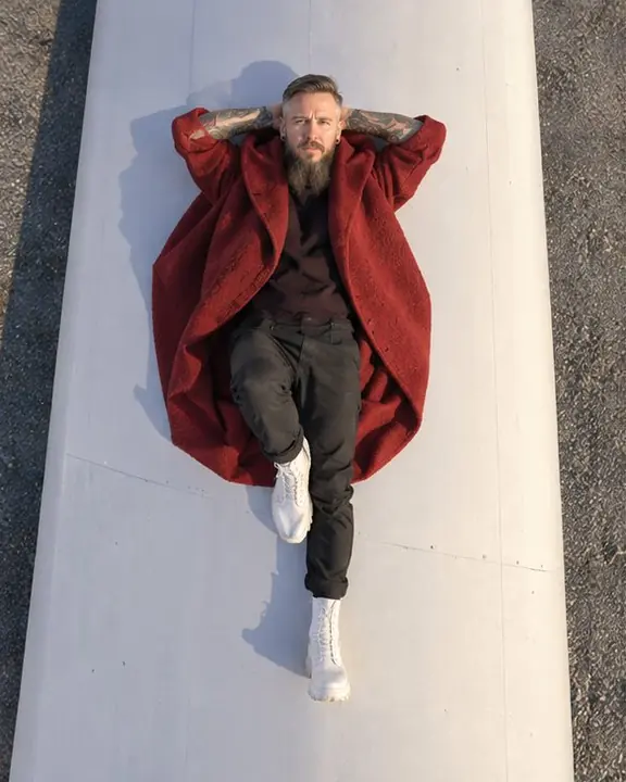 Straight-down drone view of a man in a deep red coat lying on a giant white turbine blade, looking up