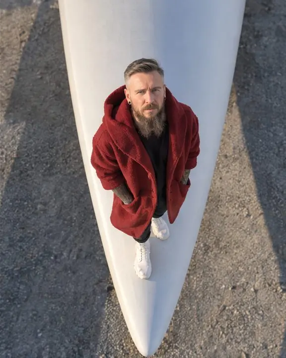 Lower drone shot of a bearded man standing at the tip of a white turbine blade, his face turned up to the camera