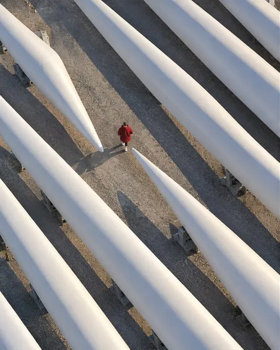 Top-down view of a man as a tiny red figure among curved white turbine blades and long blue-gray shadows