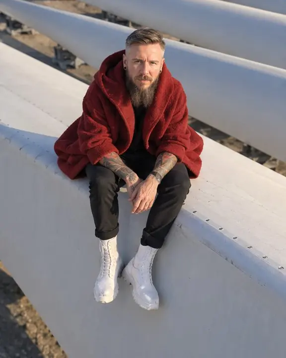A bearded man in a red cocoon coat sits on a blade edge, legs dangling, facing a low-flying drone